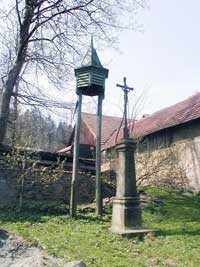 litinový křížek s kamenným podstavcem, v pozadí zvonička v Javoří